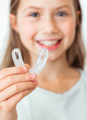 Young child with long hair smiling holding up clear retainer.