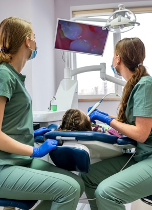 Two persons, likely dental professionals, examine a child in a dental clinic. A monitor screen displays a dental image. The room has medical equipment.