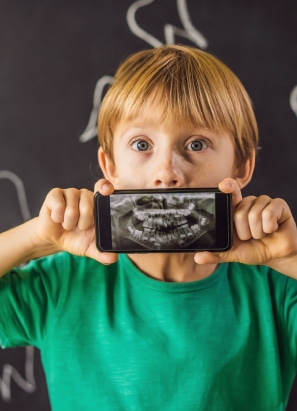 A child in a green shirt holds a phone displaying a dental X-ray in front of their mouth, against a dark background with white chalk drawings.