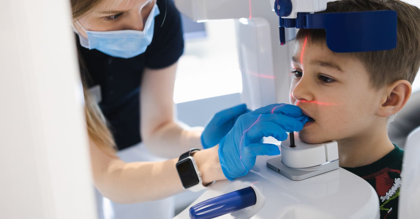 A person assists a boy with a dental X-ray machine, ensuring proper positioning and use, while wearing protective face masks and gloves.