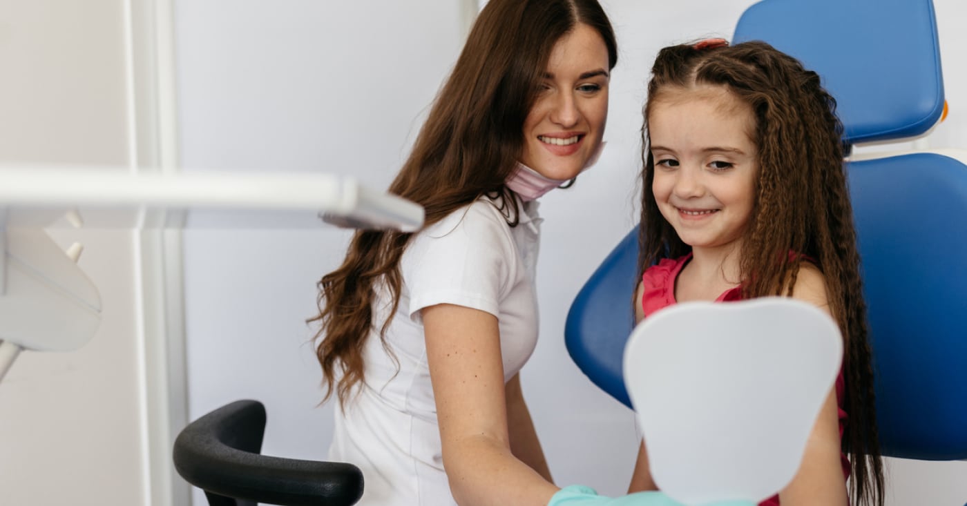 A person and a child smile in a dental clinic, with the child holding a dental mirror while sitting in a chair.