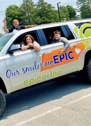 Three people interact with a branded car from "epicortho.com" displaying "Our smiles are EPIC" with an orange state outline, in a parking lot setting.