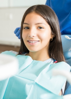 A young person with braces sits in a dental chair, wearing a blue dental bib. A person in the background wears blue gloves. No landmarks visible.