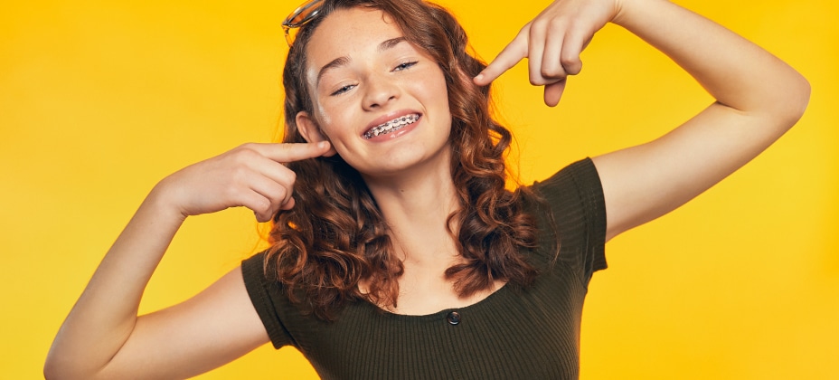 A person smiling with braces, pointing to their teeth, in front of a solid yellow background. The person is wearing a black shirt.