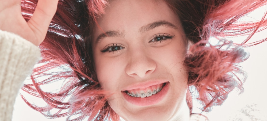 A smiling person with red hair and braces looks directly into the camera, against a bright background, creating an uplifting, cheerful atmosphere.