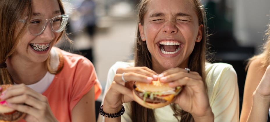 Two people are laughing joyfully while holding burgers. They appear to be outdoors in a casual setting. No landmarks or historical buildings are visible.