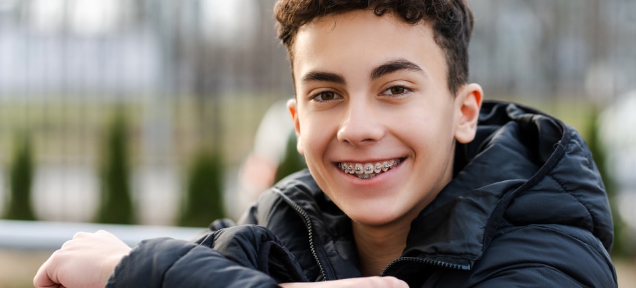 A smiling youth outdoors, wearing a black jacket, braces visible on teeth, green plants and blurry fence in the background. No recognizable landmarks.