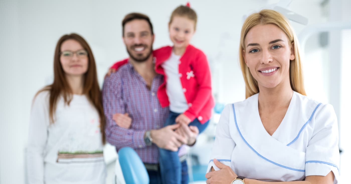 A person in a white medical uniform stands smiling, while a family of three stands in the background inside a modern, bright room.