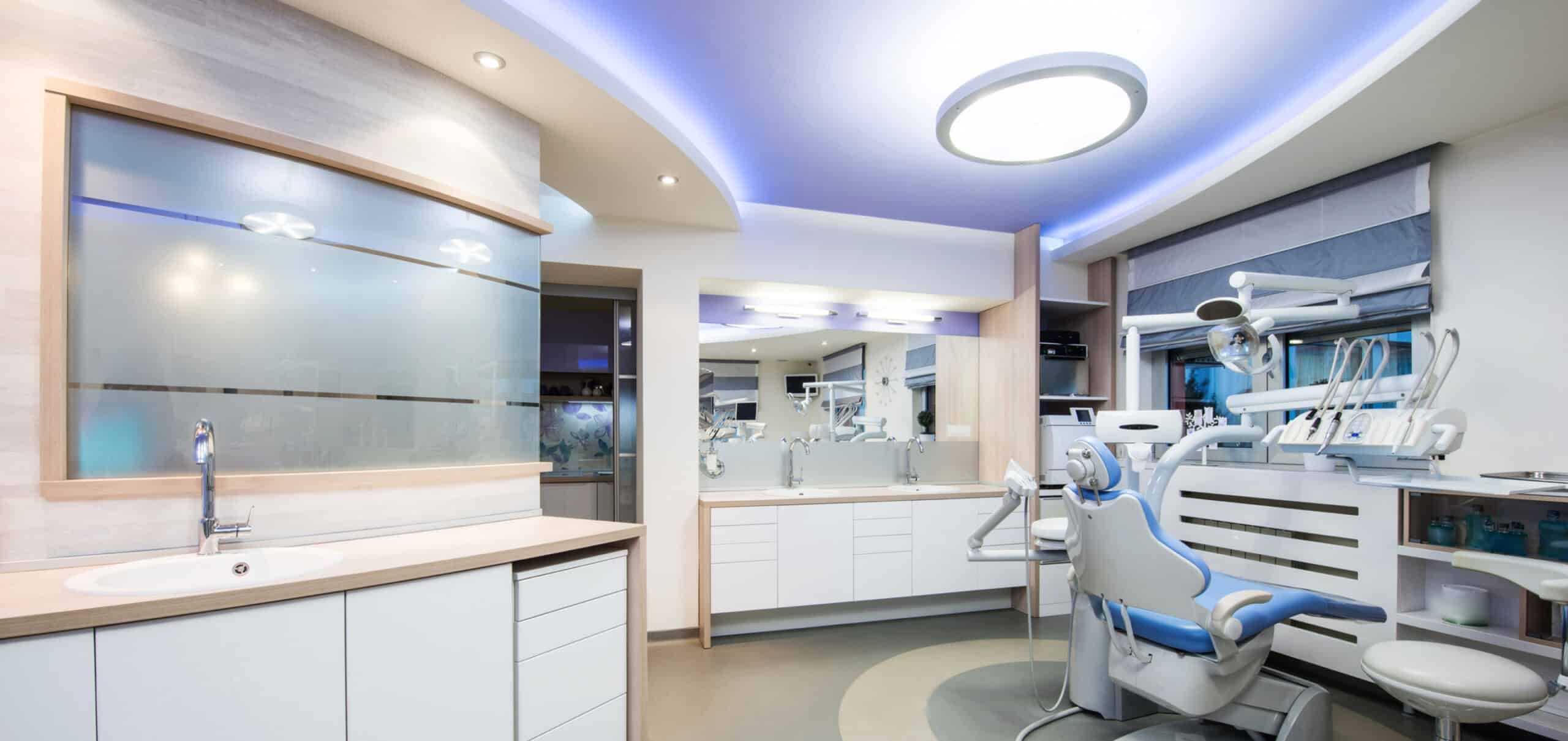 A modern dental clinic with sleek white cabinets, a dental chair, and frosted glass partitions, illuminated by soft blue and white lighting.