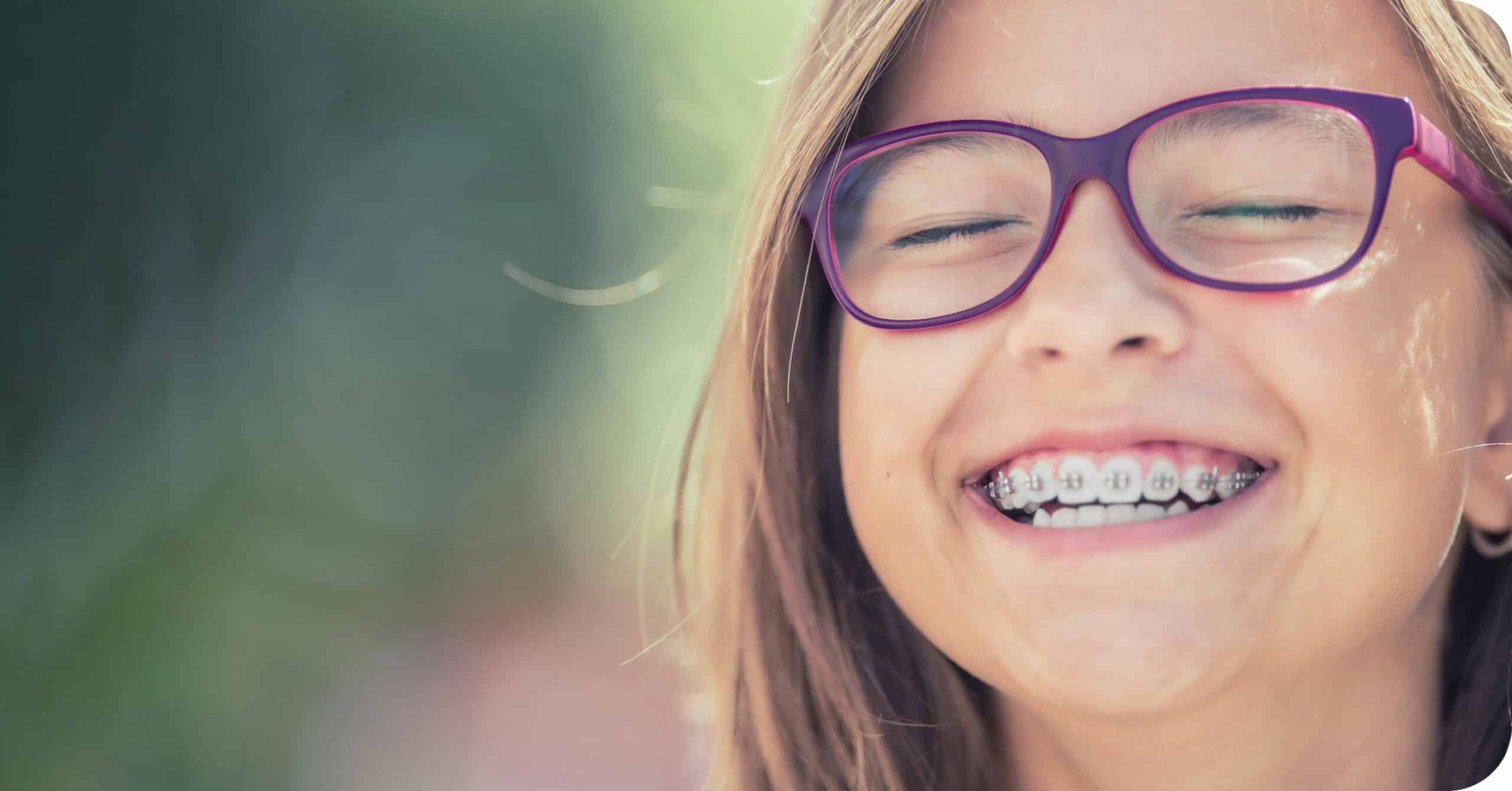 A child wearing glasses and braces smiles broadly. The background is blurred with green foliage, indicating an outdoor setting.