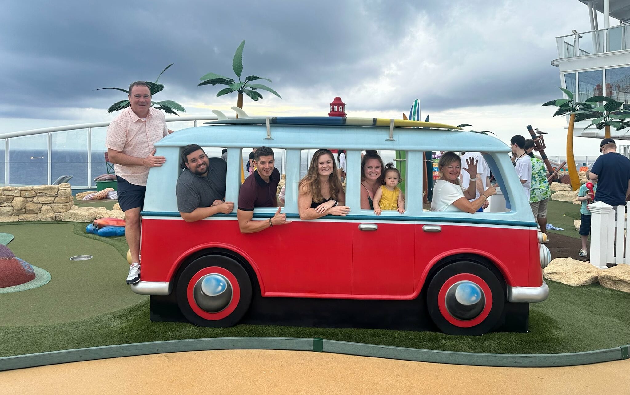 Several people pose inside and around a red cartoon-style van installation, with artificial palm trees and a cloudy sky in the background.