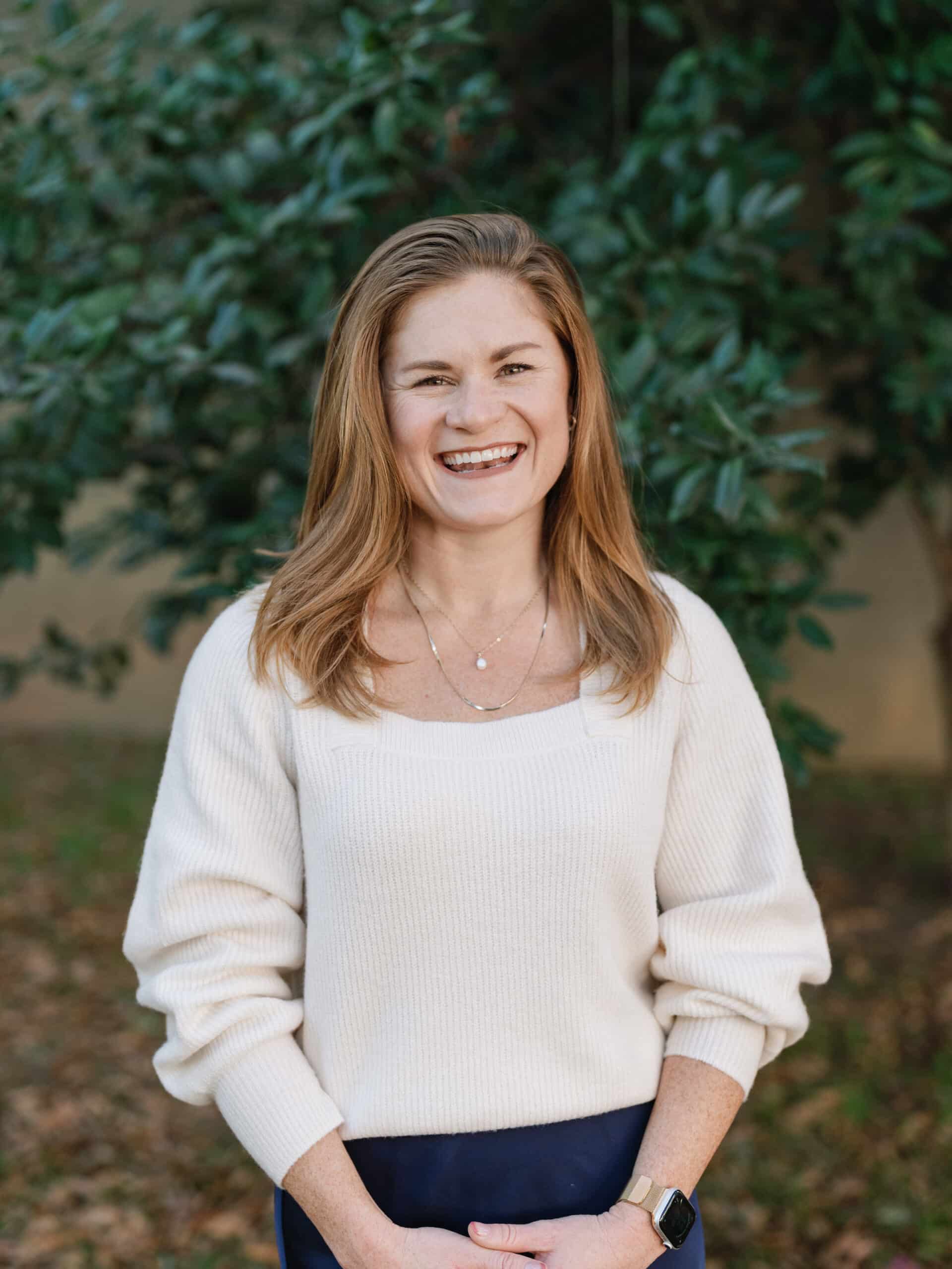 A person wearing a white sweater smiles in front of green foliage, hands clasped. The background features trees and shrubbery.