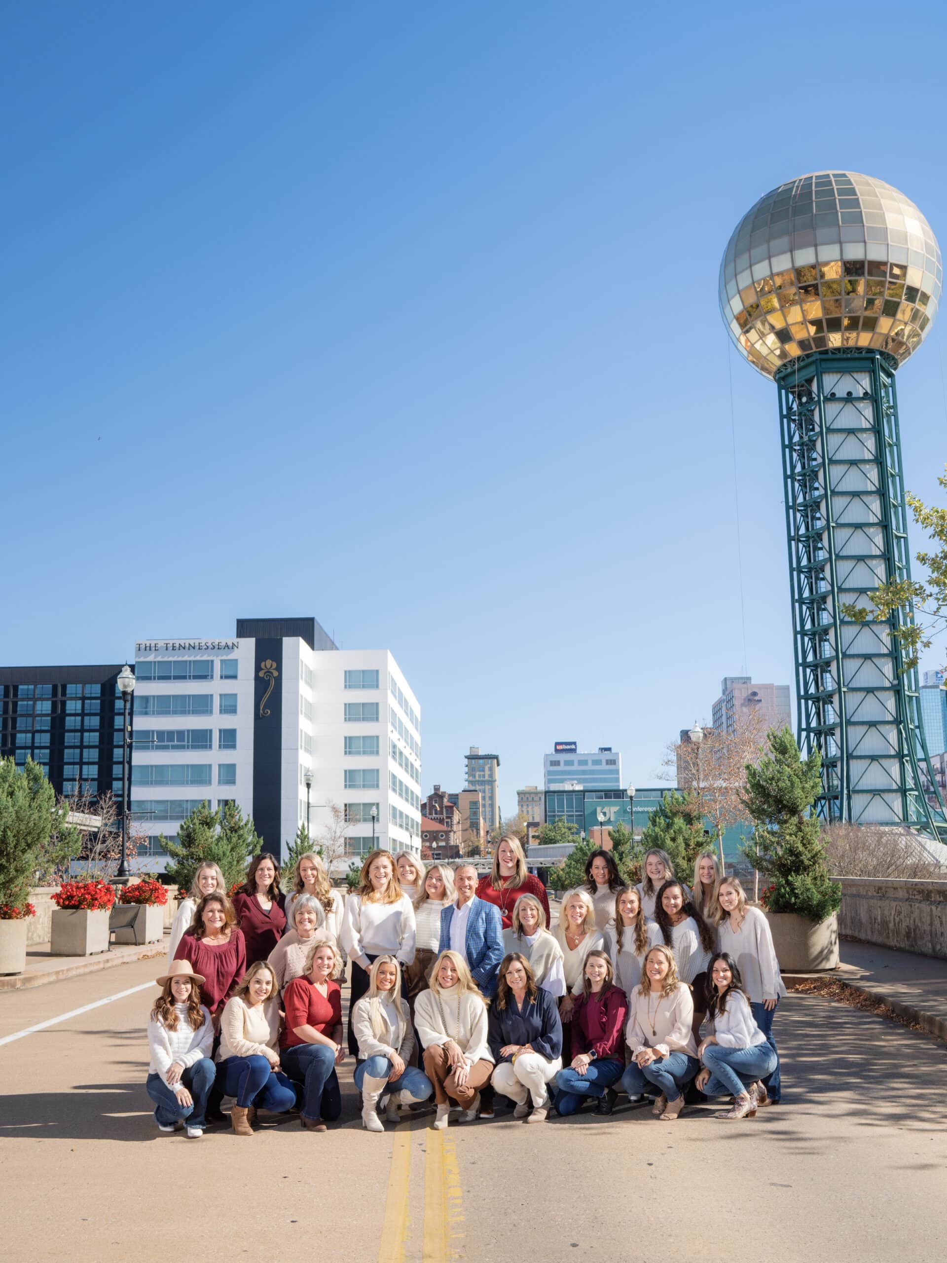 A group of people poses in front of the Sunsphere in Knoxville, with modern buildings and clear blue sky in the background.