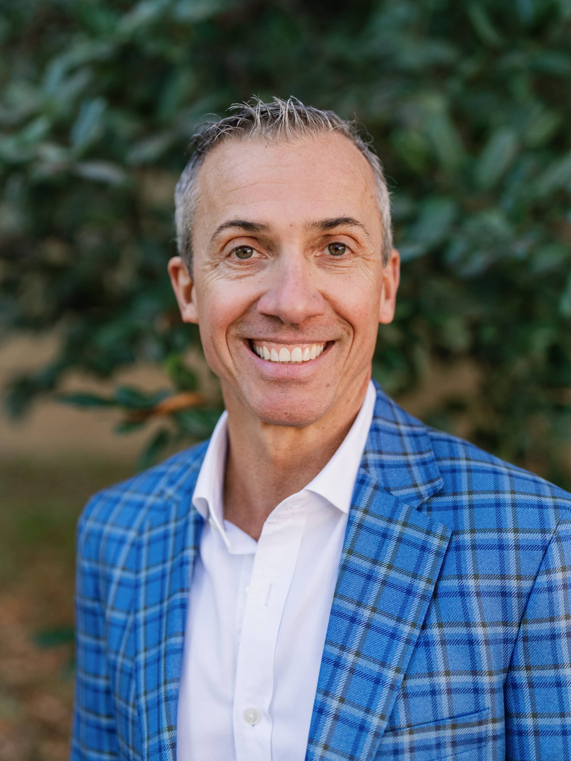 A smiling person in a blue plaid jacket, standing outdoors with greenery in the background. No recognizable landmarks are present.