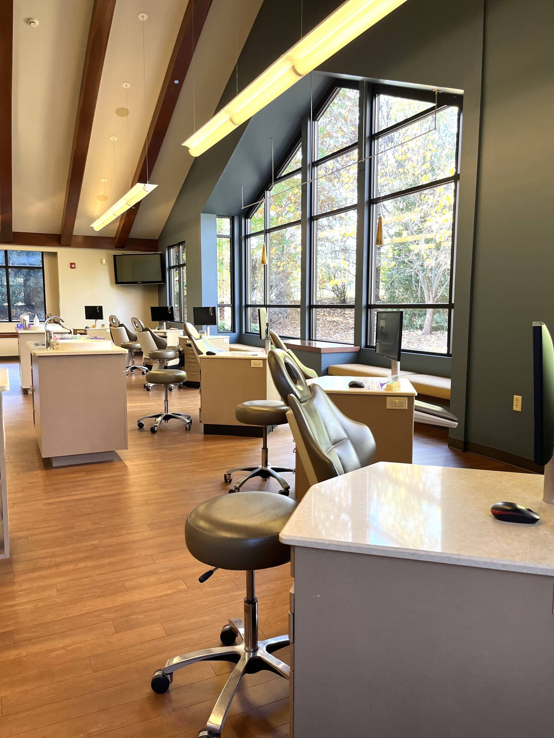 A modern dental office with rows of dental chairs and workstations, featuring large windows and wooden floors, overlooking a scenic outdoor view.