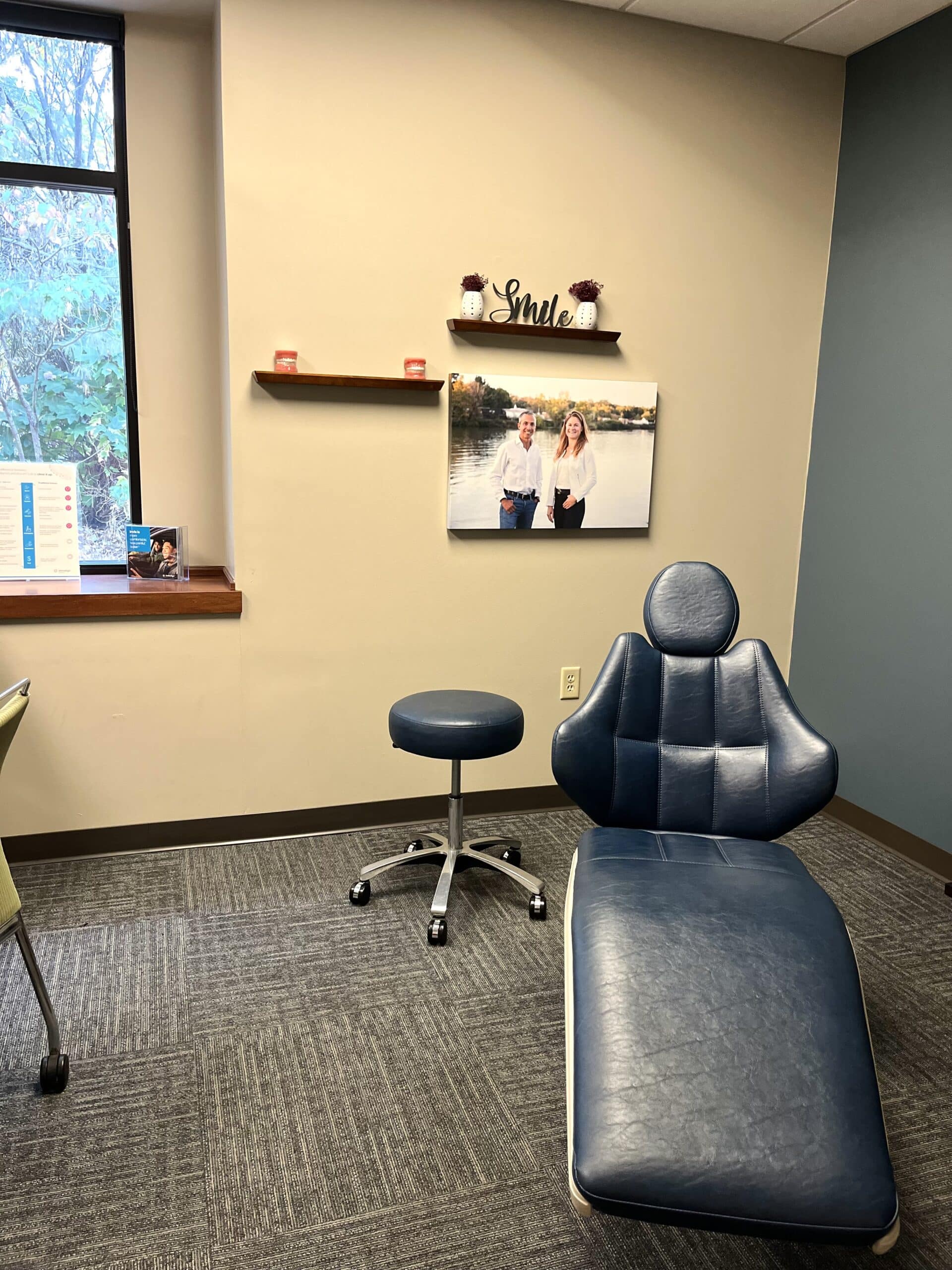 Dental office with a blue chair, stool, and window. Photograph of two people on the wall, shelf with "Smile" decoration above.