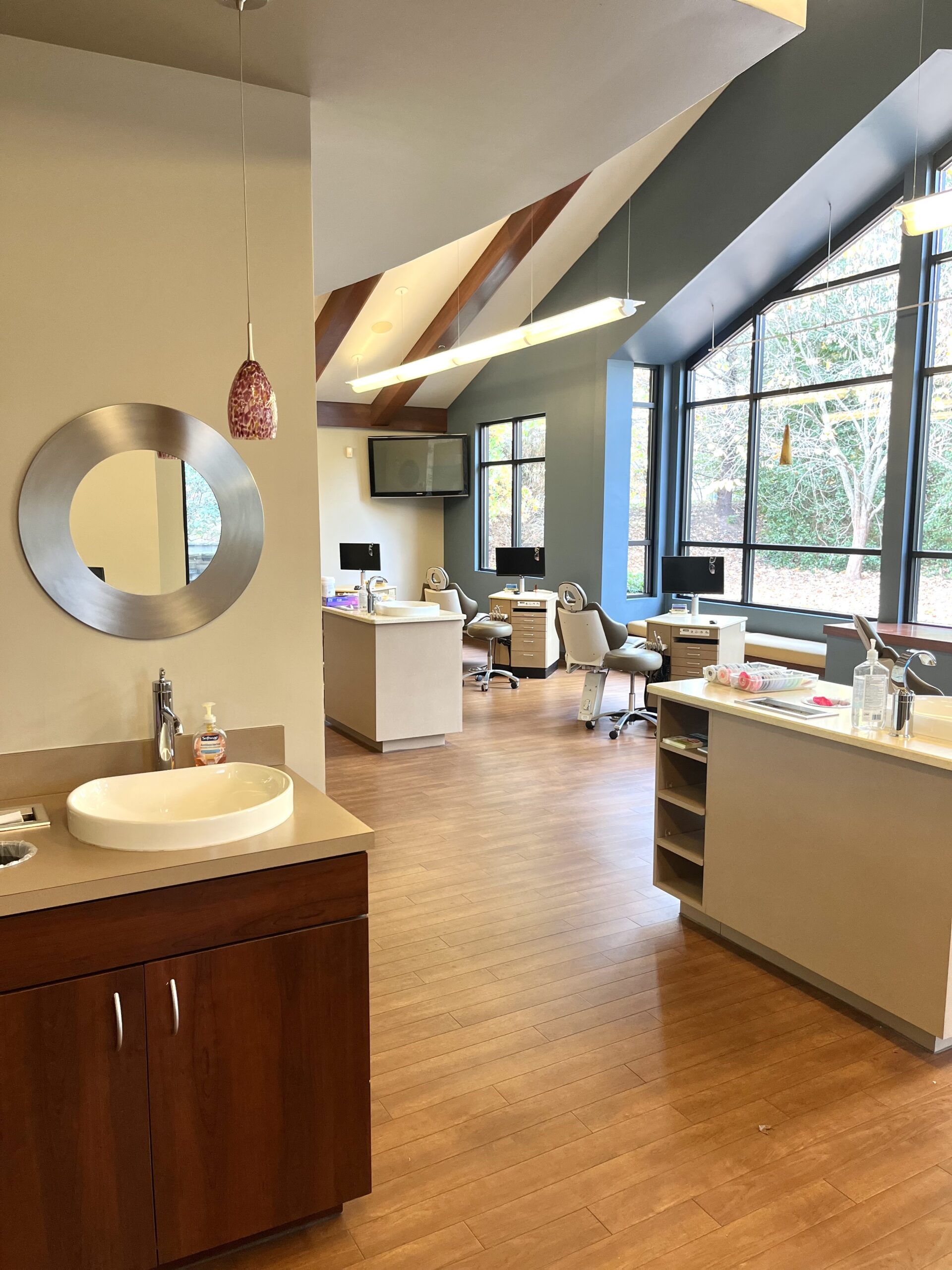 Modern dental office interior with wooden flooring, workstations, and large windows. Mirrors, sinks, and equipment are visible. Bright and welcoming atmosphere.