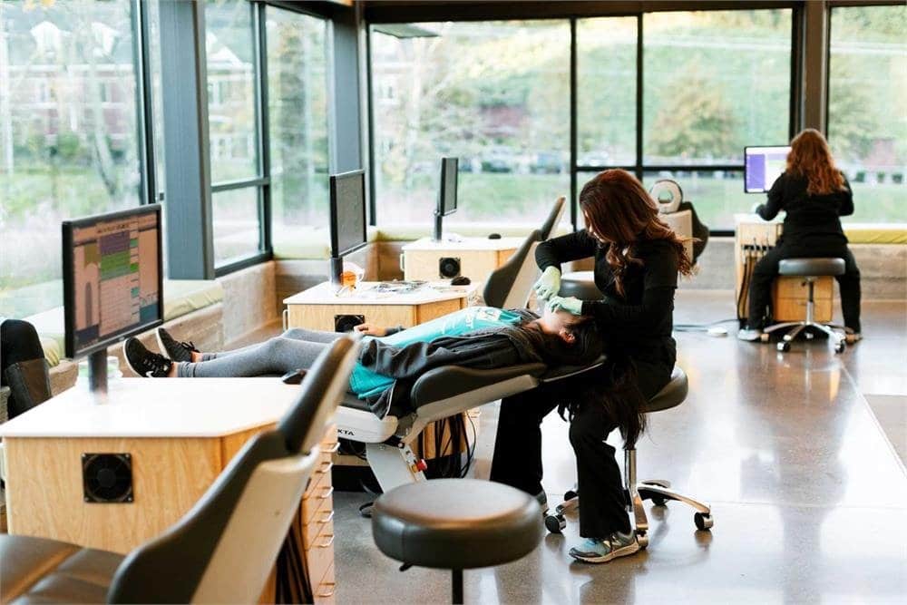 A modern dental clinic features a person receiving treatment in a reclining chair, while another works at a computer in a well-lit room.