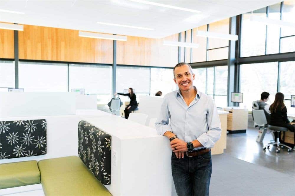 A person stands smiling in a modern office with others working at computers. Bright, spacious interior with natural light and wooden accents.