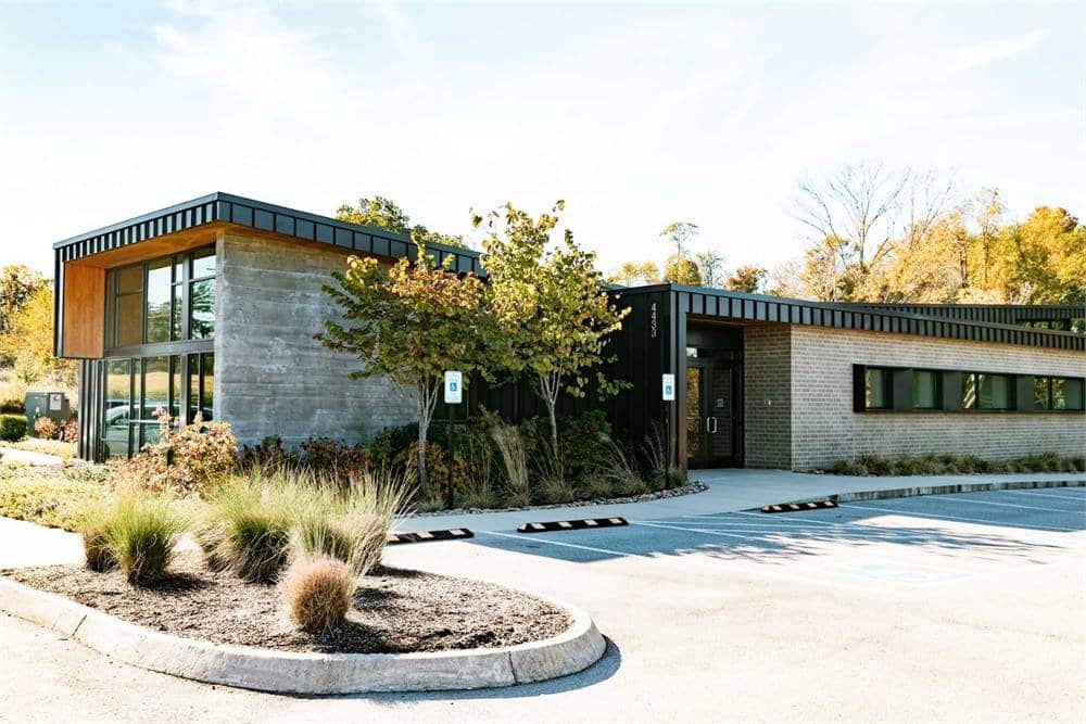 A modern, single-story building with large windows is surrounded by trees and landscaping in a quiet, sunny setting. No people are present.