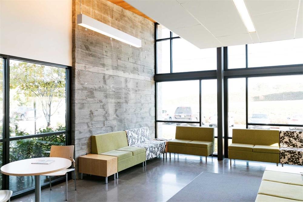 Modern waiting area with large windows, green and beige seating, a round table, and concrete walls. No people or recognizable landmarks are visible.