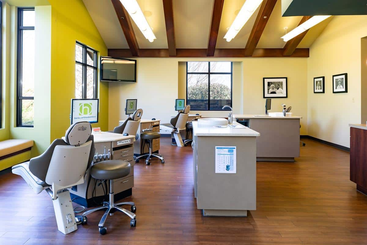 Bright dental office with multiple treatment stations, computers, and dental chairs. Windows and framed photos on walls. Modern, clean, and welcoming atmosphere.