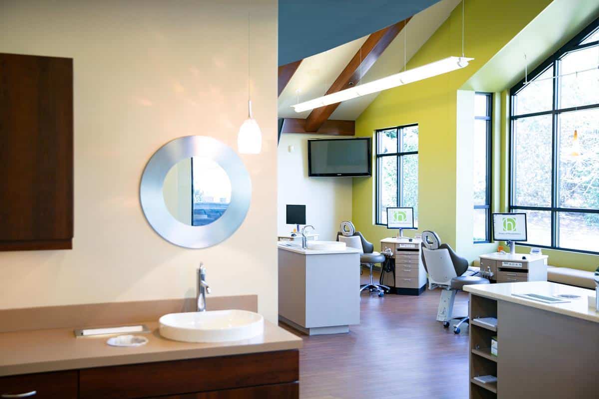 Modern dental office interior with bright yellow walls, large windows, computer stations, and a circular mirror near a sink. Peaceful and inviting atmosphere.