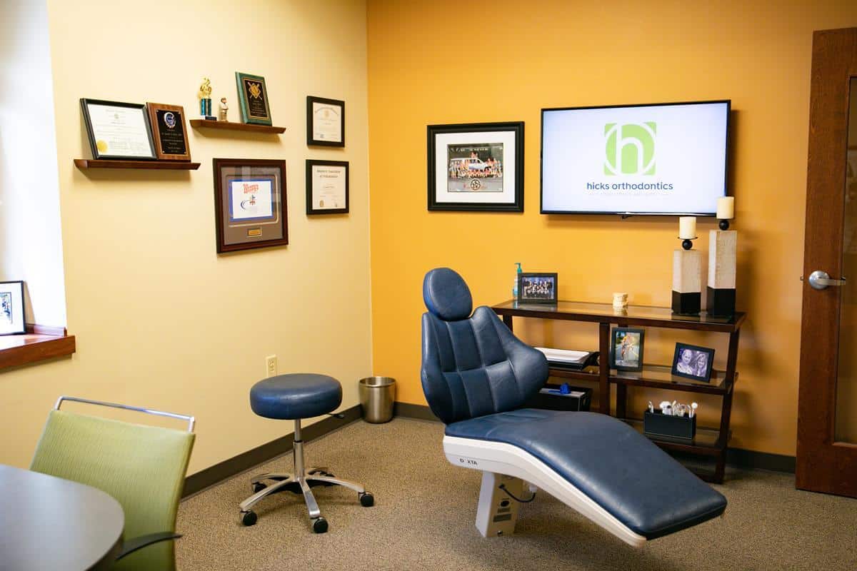 An orthodontic office with a dental chair, framed certificates on the wall, shelves with decor, and a screen displaying "Hicks Orthodontics."