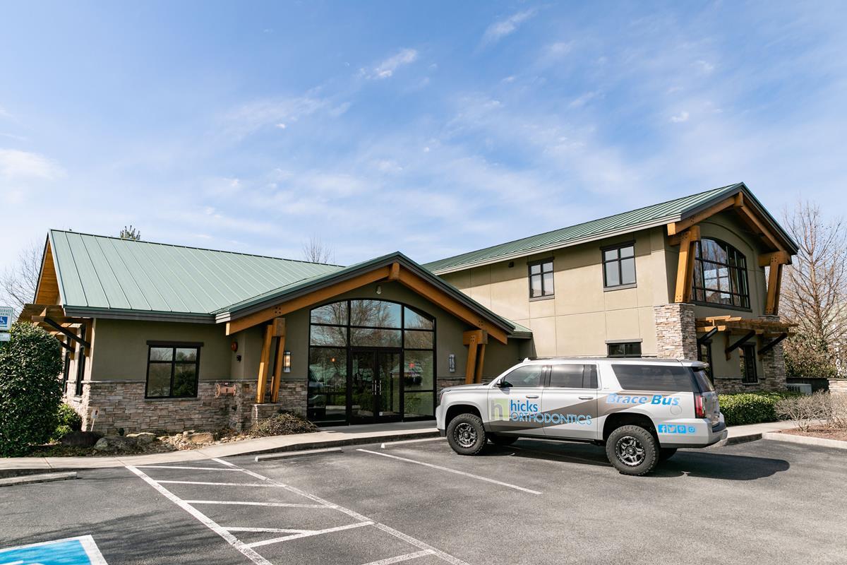 A modern building with a green roof and large windows is seen in a parking lot with a branded vehicle parked nearby.