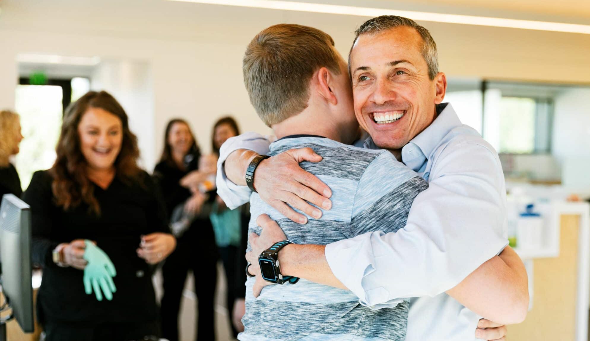 Two people embrace in an office setting while others smile in the background. The atmosphere is cheerful and lively.