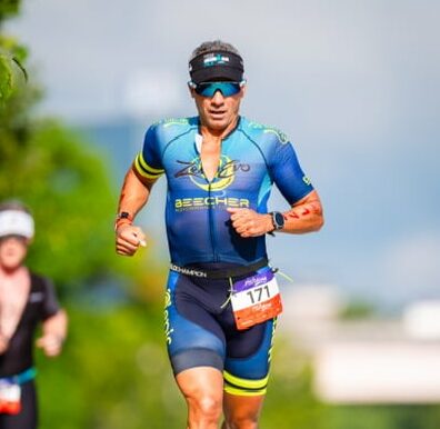 A person in a triathlon suit runs outdoors during a race, with another runner in the background. No recognizable landmarks are visible.