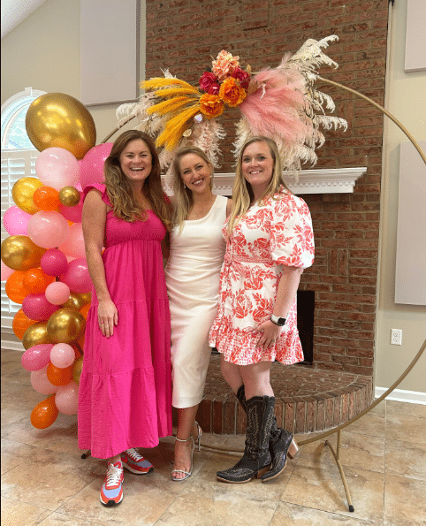 Three people smile in front of a decorative brick fireplace, with colorful balloons and floral arrangements enhancing the background.