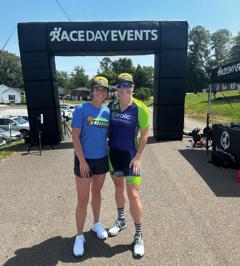 Two people stand under a "Race Day Events" arch, wearing athletic gear. Trees and parked cars are visible in the background.