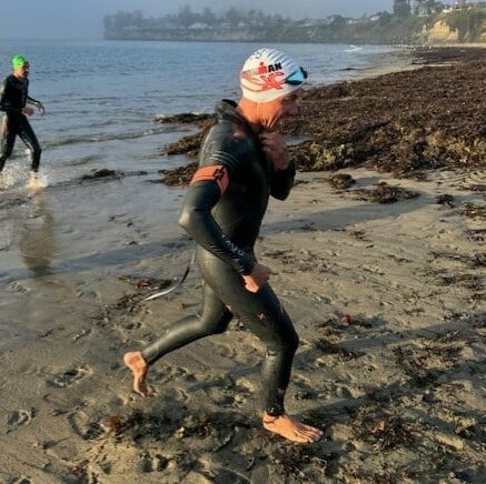 Two people in wetsuits and swim caps exit the water onto a sandy beach, with cliffs and houses visible in the background.