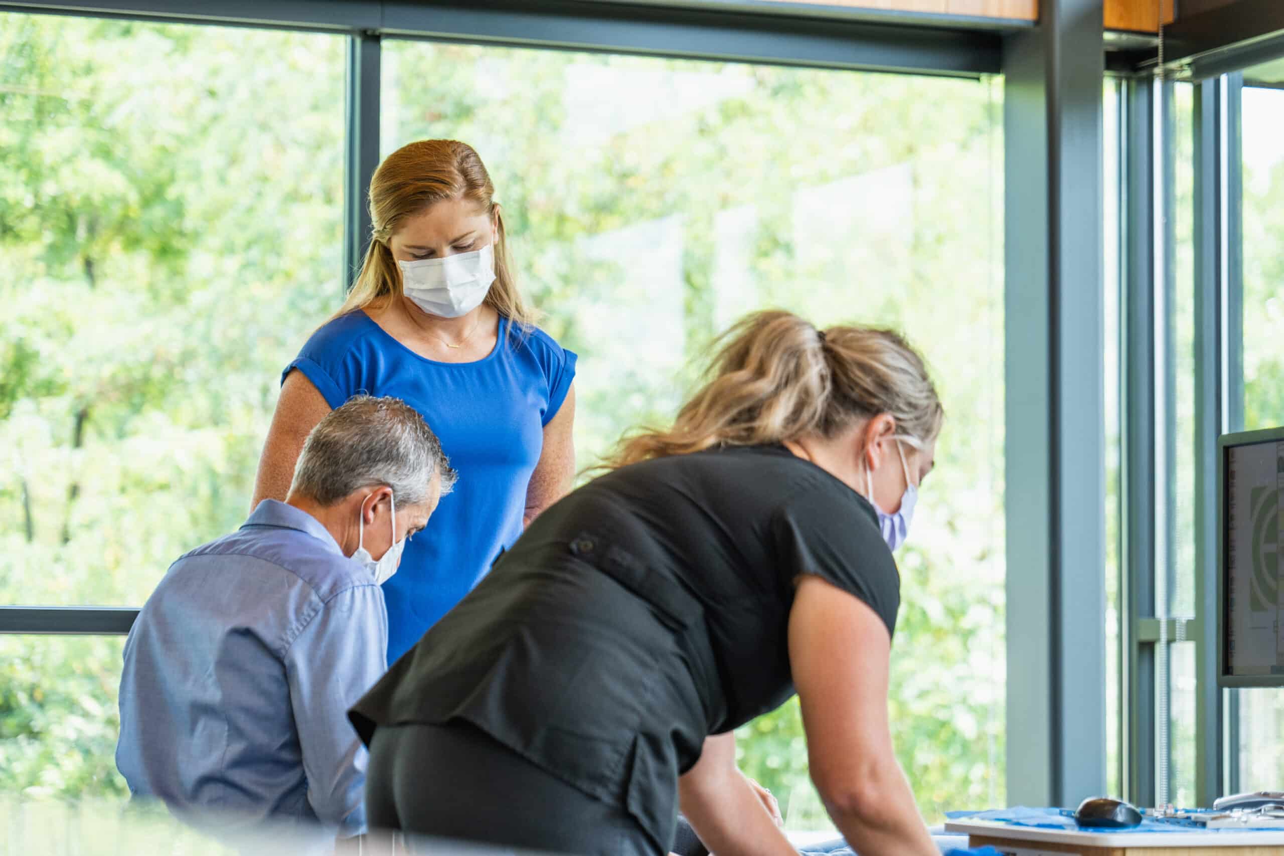 Three people wearing masks are collaborating in a modern office with large windows overlooking greenery. They are focused on work materials and a computer.