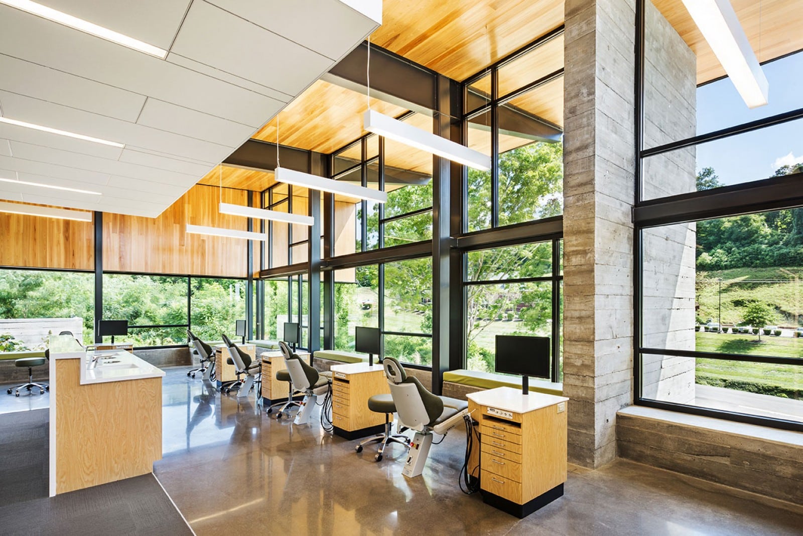 Modern office interior with large windows, overlooking lush greenery. Open workspace features wooden elements, sleek desks, and computer stations. Bright natural light fills the room.