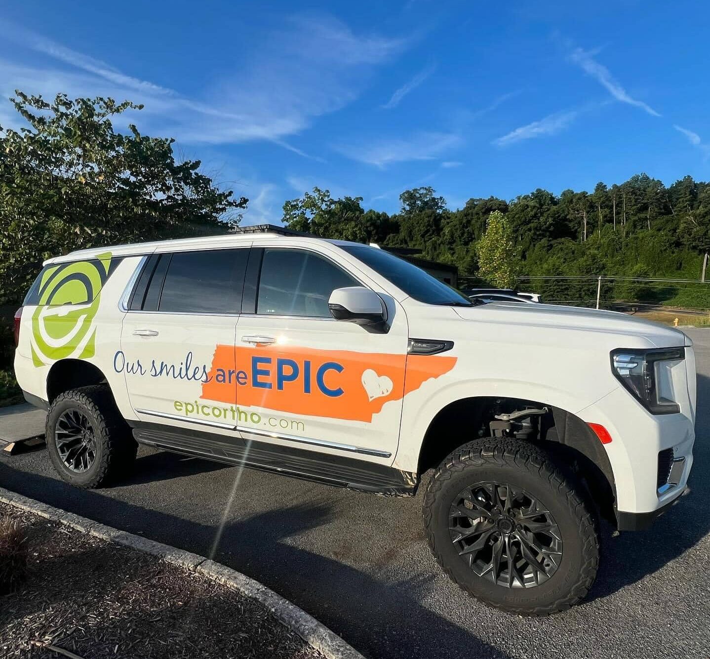 A branded SUV with "Our smiles are EPIC" parked on a sunny day, surrounded by greenery and a clear blue sky.